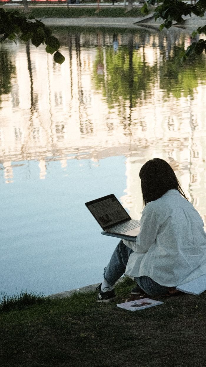 A woman working remotely on a laptop by a pond in a peaceful park setting, reflecting calm urban architecture.