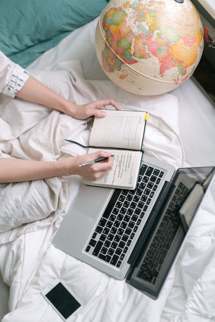 Overhead view of a cozy workspace with a laptop, notebook, and globe on a bed.